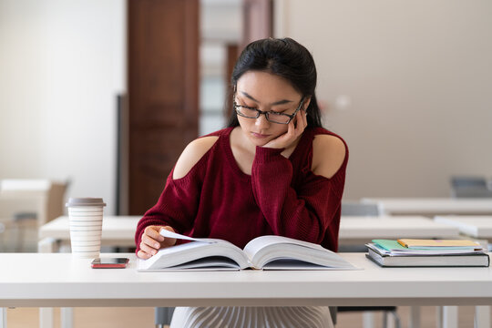 Geeky nerdy asian girl in glasses sitting at desk in library reading book, studying in reading room, female student using textbook preparing for exam or test, selective focus. Education concept