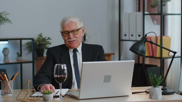 Senior man freelancer drinking wine from glass after working on laptop computer pc sitting at desk at home office. Elderly mature guy e-learning, online distant video call conversation remote job