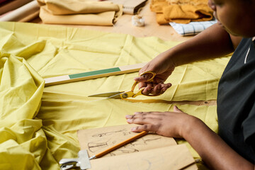 Young woman with notebook of sketches and scissors cutting yellow fabric while working over new...