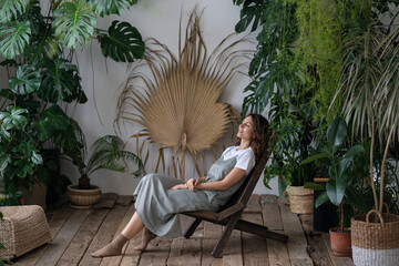 Young calm hipster woman relaxing sit on comfortable armchair in stylish scandinavian indoor garden...