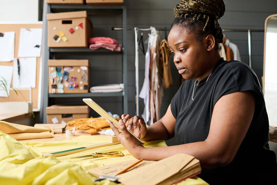 Young serious female tailor with tablet looking through new online orders of clients while sitting by workplace with textile and lookbook
