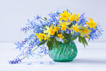 Bouquet of spring flowers in a vase on a white wooden table. Yellow daffodils, blue scilla, muscari, anemones. Festive garden still life, blur. - obrazy, fototapety, plakaty
