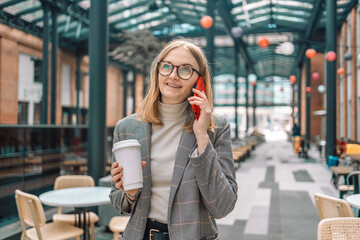 Fototapeta premium Happy beautiful businesswoman talking on cell phone outside, standing with coffee paper cup near coffee shop outdoors while walking in morning city. 