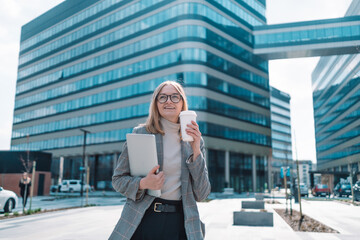 European blonde attractive business woman in trendy clothes with a laptop and take away Bio paper coffee cup. Eco material. Glass for hot drinks. Empty space for text, mock up