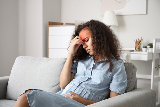 Pregnant African-American woman with headache sitting on sofa at home