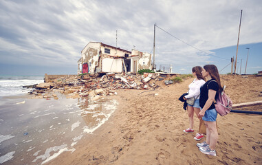 Two women watching how the high tide created by a tsunami destroys the houses located on the coast, in front of a beach. Ruined houses are reduced to rubble and disappear with the waves of the ocean. - obrazy, fototapety, plakaty