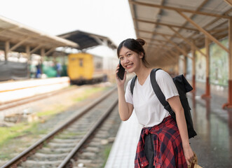 Beautiful young asian woman with a backpack uses the phone while standing near the railroad train...