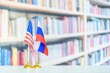 Flag of USA and Russian federation on a table over a blurred bookshelf background. A symbol of...