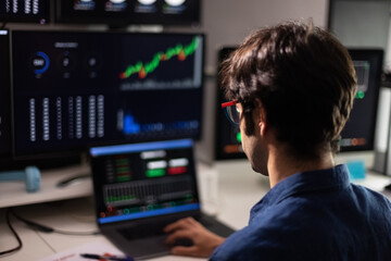 Back view of man in blue shirt working with multiple screens and laptop. Stock market and trading.