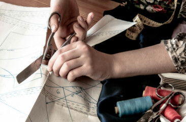 Obraz premium The girl cuts the fabric with scissors. In the background is a sewing machine. The fabric is white and pink. Sewing content.