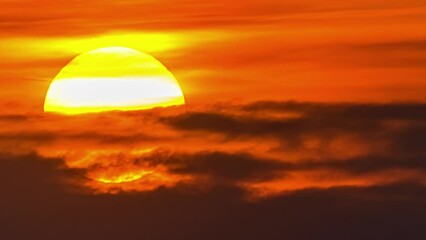 View of yellow sun setting in the orange sky with dark clouds passing by in timelapse. Close up view of big round yellow sun going down over orange sky.