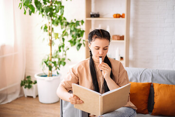 A beautiful young woman uses a disposable electronic cigarette on the couch at home reading a book....