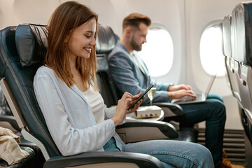 Cheerful woman using mobile phone in airplane - obrazy, fototapety, plakaty