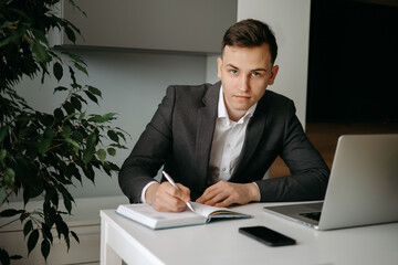 Serious young Caucasian man sitting at desk making notes in notebook