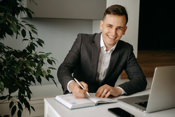 Naklejka premium Happy young Caucasian man sitting at desk and taking notes in notebook