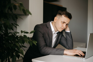 Thoughtful young businessman looks at the monitor. Serious young Caucasian man sit at desk at home office work on laptop online with notebook