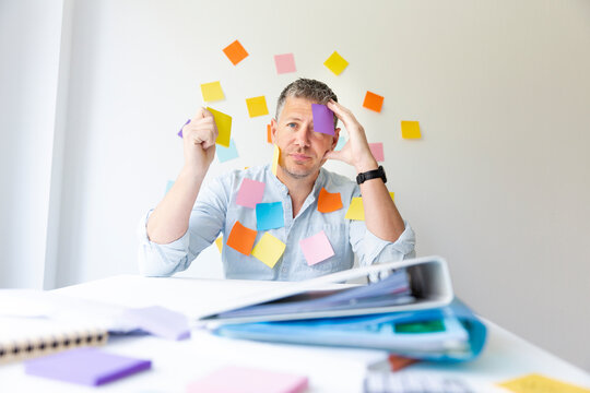 Man sits behind white office desk in front of wall with many colorful notes and also has on himself stick many notes and is looking for an idea