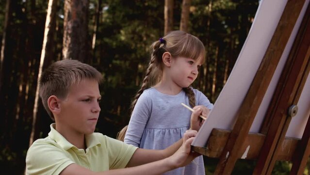 Happy family boy and girl drawing coloring picture with brush helping cute child daughter enjoying talk play children, mom dad and kid girl having fun in park. Family walk green grass in natural park