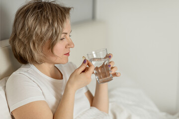 a middle-aged white woman with moisturized beautiful skin holds a glass of clean water and vitamins in her hands while sitting on the bed. the concept of health and beauty care