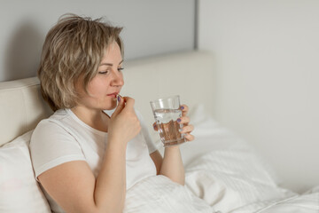 a middle-aged white woman with moisturized beautiful skin holds a glass of clean water and vitamins in her hands while sitting on the bed. the concept of health and beauty care