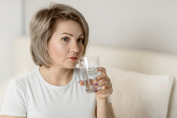 a middle-aged white woman with moisturized beautiful skin holds a glass of clean water in her hands, sitting on the bed. the concept of health and beauty care