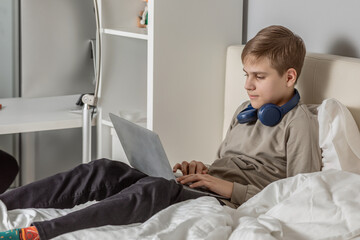 Obraz premium a teenage boy is sitting on a bed with a laptop in his hands and headphones around his neck, working on a laptop