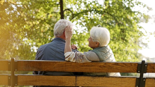 Outdoor photo of senior caucasian heterosexual couple sitting on a bench in the park enjoying beautiful weather. Gray-haired lady in warm vest touches her husband's hair. High quality photo