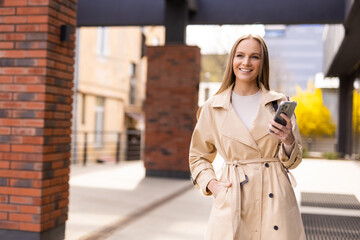 Young pretty woman in an autumn coat uses a smartphone in a European city.