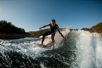 athletic man in wetsuit on wakesurf board riding down the splashing wave