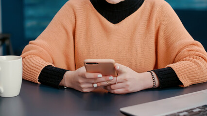 Cheerful woman browsing internet on smartphone at desk, doing college research and attending online lesson course. Young adult working on mobile phone to plan educational research project.