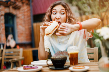 A girl in a hurry bites a burger on the run in a fast food cafe and looks at the smart watch. Late...