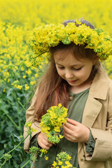 a cute six-year-old girl in a khaki cotton dress stands in a rapeseed field with a wreath of rapeseed flowers on her head