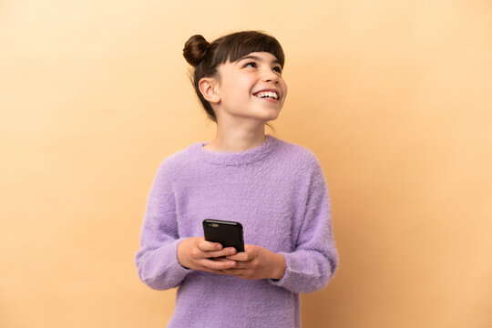 Little caucasian girl isolated on beige background using mobile phone and looking up