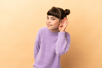 Little caucasian girl isolated on beige background listening to something by putting hand on the ear