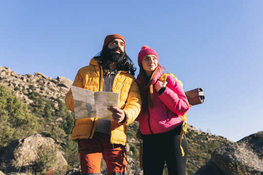Bearded man with map standing by girlfriend wearing backpack on sunny day