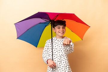 Little girl holding an umbrella isolated on beige background inviting to come with hand. Happy that you came