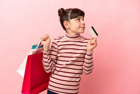 Little caucasian girl isolated on pink background holding shopping bags and a credit card and thinking