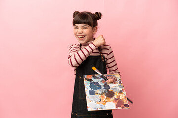 Little artist girl holding a palette isolated on pink background celebrating a victory
