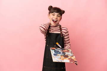 Little artist girl holding a palette isolated on pink background laughing