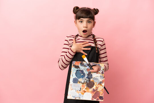 Little artist girl holding a palette isolated on pink background surprised and shocked while looking right