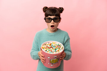 Little caucasian girl isolated on pink background surprised with 3d glasses and holding a big bucket of popcorns