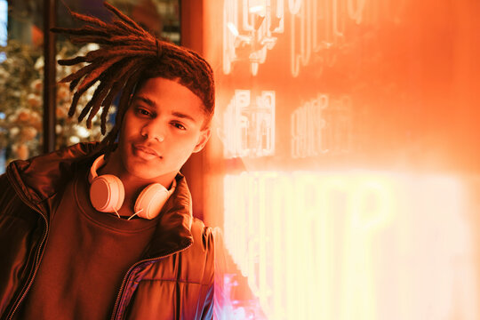 Young man wearing wireless headphones leaning on illuminated wall at night