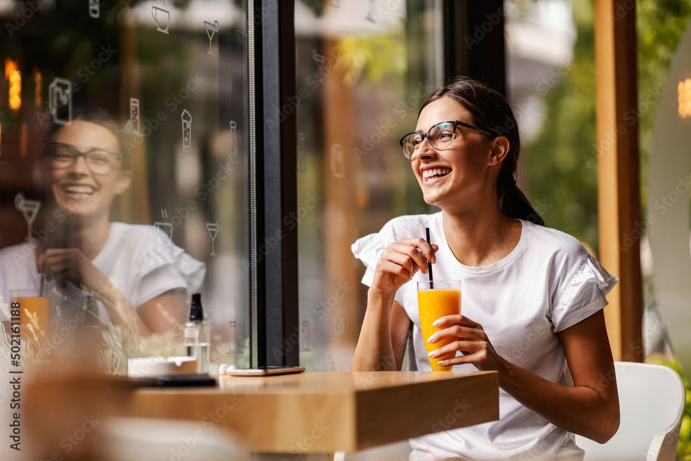 Wall mural a happy young woman is sitting in a restaurant and drinking fresh orange juice instead of coffee.