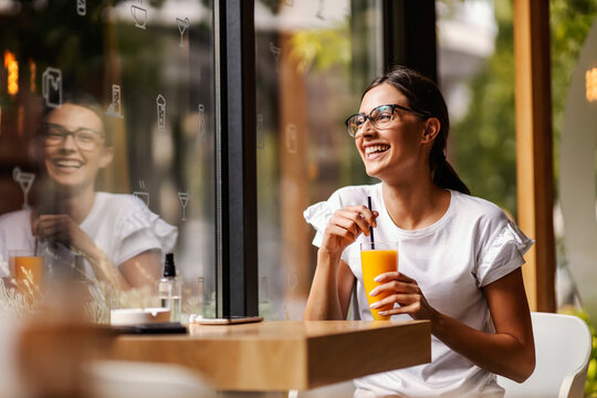 A happy young woman is sitting in a restaurant and drinking fresh orange juice instead of coffee. 