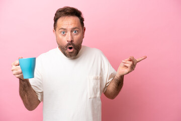 Middle age caucasian man holding cup of coffee isolated on pink background surprised and pointing side