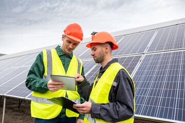 two strong skilled men in helmets and vests stand and talk in solar panels.