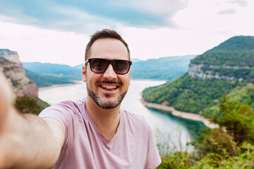 Young cheerful handsome man with beard, wears sunglasses taking selfie on top of mountain lake - Adventure and travel lifestyle concept.