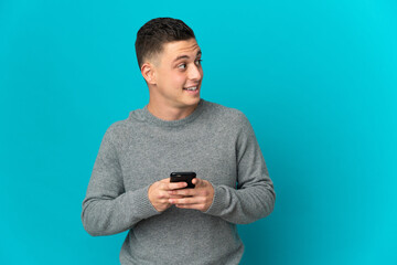 Young caucasian man isolated on blue background using mobile phone and looking up