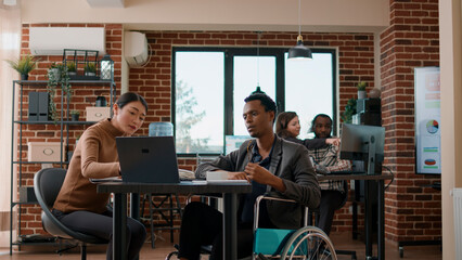 African american man with handicap talking to colleague about business growth, analyzing financial charts to plan development. People brainstorming ideas in didability friendly office.
