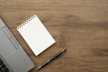 Blank notebook with pen are on top of wood office desk table with laptop computer. Top view with copy space, flat lay.